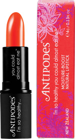 Antipodes Piha Beach Tangerine Lipstick in a black tube beside its box with sunset and beach illustrations.