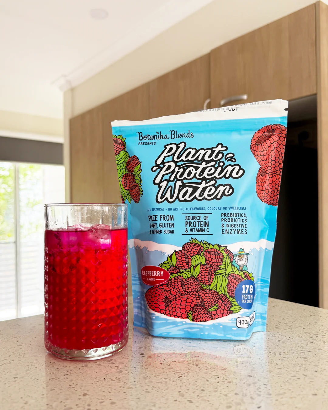 Botanika Blends Raspberry Plant Protein Water served in a glass with fresh raspberries and a 400g tub.
