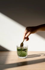A glass of green superfood drink next to an Ora Organic Greens sachet, surrounded by fresh organic ingredients.