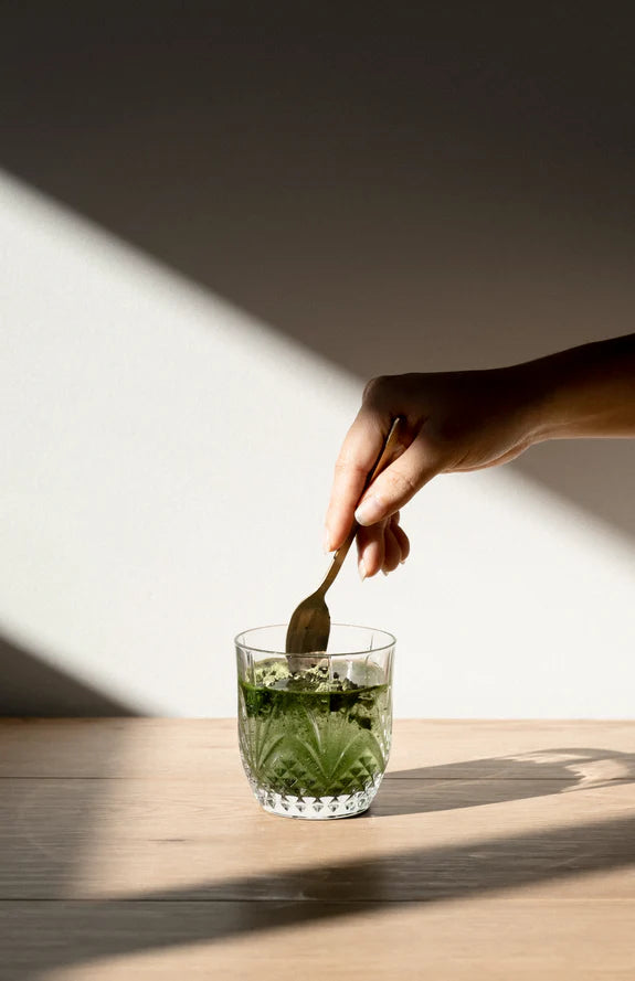 A glass of green superfood drink next to an Ora Organic Greens sachet, surrounded by fresh organic ingredients.