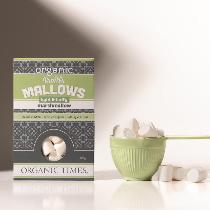 A bowl of white Organic Times Vanilla Mallows marshmallows with two mugs of hot chocolate on a wooden surface.